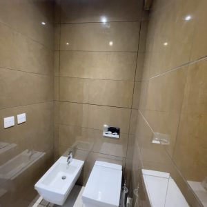 A compact bathroom with beige tiled walls, featuring a wall-mounted toilet and bidet. The hinged tile door access panel is closed, blending into the wall.