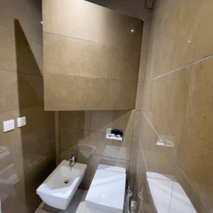A bathroom with a wall-mounted toilet and bidet. The hinged tile door access panel on the wall is partially open, exposing the concealed space behind it.