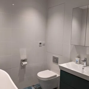 A contemporary bathroom with white tiled walls, a floating toilet, and a modern sink vanity. A hidden hinged tile door access panel is flush with the wall, maintaining the clean design.