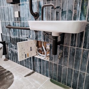A contemporary bathroom with white tiled walls, a floating toilet, and a modern sink vanity. A hidden hinged tile door access panel is flush with the wall, maintaining the clean design.