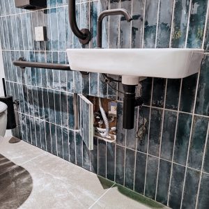A sleek bathroom with a hinged tile door access panel open, revealing a storage compartment containing towels, toiletries, and cleaning supplies.
